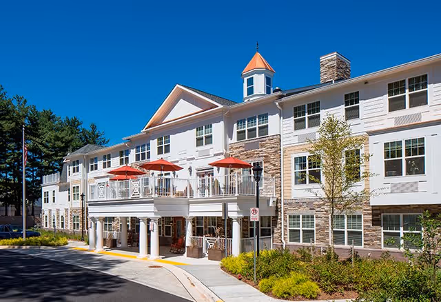 Exterior view of a senior living facility building with white and stone facade, multiple windows, a balcony with red umbrellas, and a small tower with a pointed roof. The building is surrounded by landscaped greenery and a clear blue sky is visible.