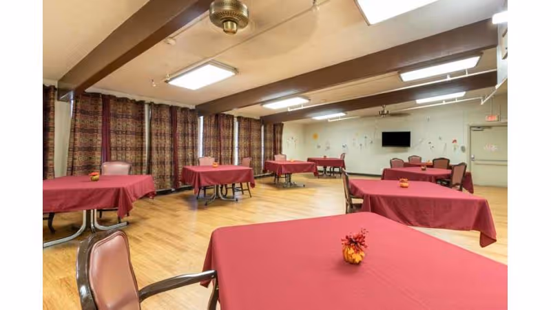 A spacious dining room with multiple tables covered in red tablecloths, each with a small decorative centerpiece. The room has wooden flooring, patterned curtains covering the windows, a ceiling fan, fluorescent lighting, and a wall-mounted television. Chairs are arranged around the tables.