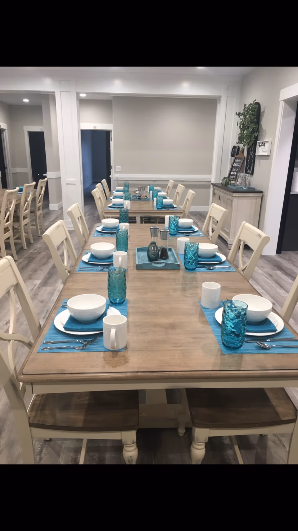 Long communal dining room with wooden tables set with teal placemats, bowls, and glasses and white chairs.