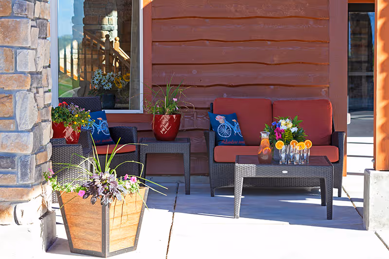 Outdoor seating area with two dark wicker chairs and a loveseat with red cushions and blue decorative pillows. A small wicker table holds a pitcher of iced tea and glasses garnished with orange slices. Several potted plants with colorful flowers are placed around the seating area, which is set against a wooden exterior wall.