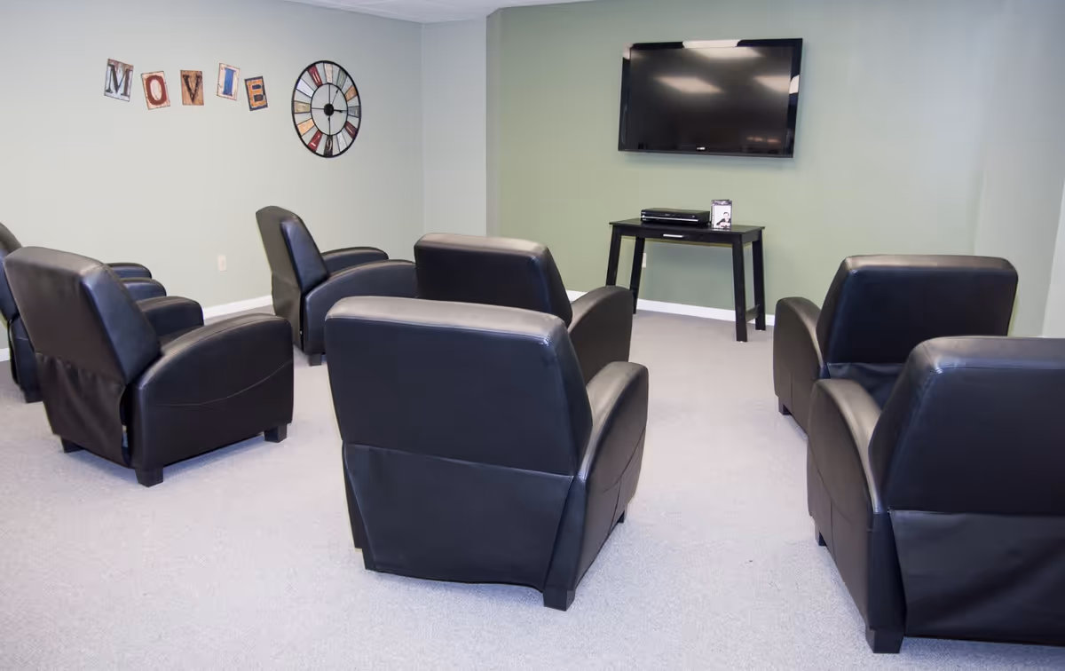 A small movie viewing room with six black leather recliners arranged in two rows facing a wall-mounted flat screen TV. A small black table beneath the TV holds a DVD player and a DVD case. The walls are painted light green, and there is a decorative clock and letters spelling 'MOVIE' on the wall.