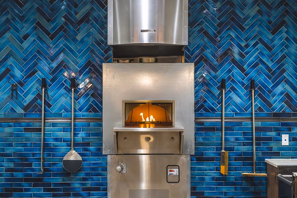A stainless steel wood-fired oven with a small fire burning inside, mounted against a wall covered with blue herringbone and rectangular tiles. Four long-handled kitchen tools hang on the wall, two on each side of the oven.