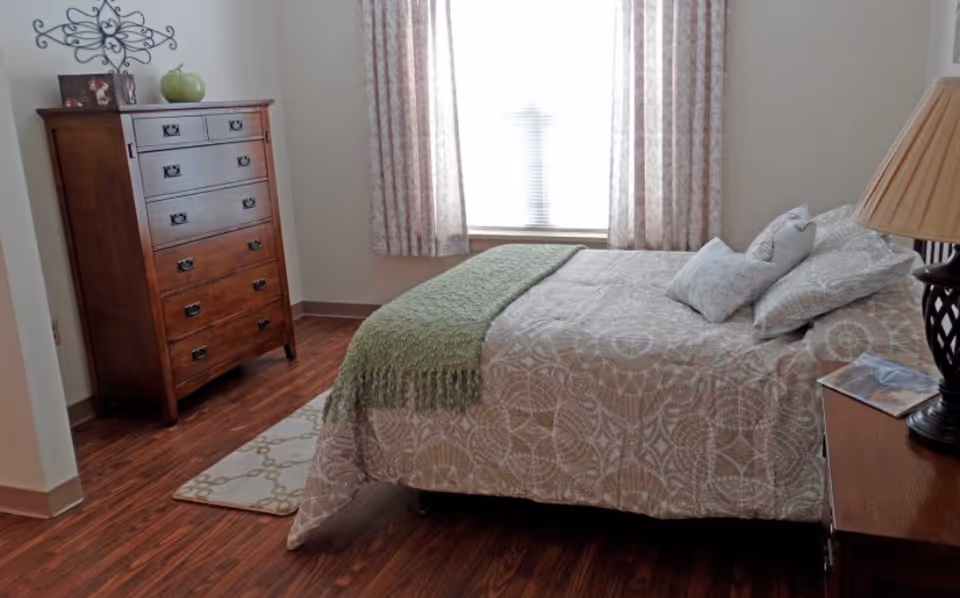 A cozy bedroom with a wooden dresser on the left, a bed with patterned bedding and multiple pillows, a green throw blanket, a bedside table with a lamp and a magazine, and a window with patterned curtains letting in natural light.