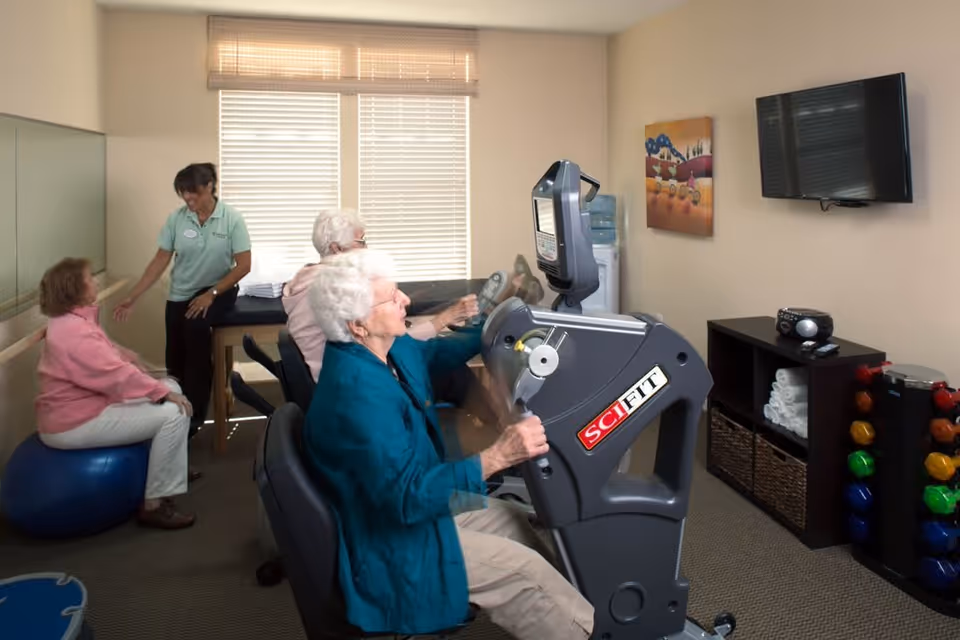 A small exercise room with elderly women using fitness equipment. One woman is seated on an exercise ball, another is using a seated arm bike machine, and a staff member is assisting. The room has a window with blinds, a wall-mounted TV, a rack with colorful dumbbells, and shelves with towels and a radio.
