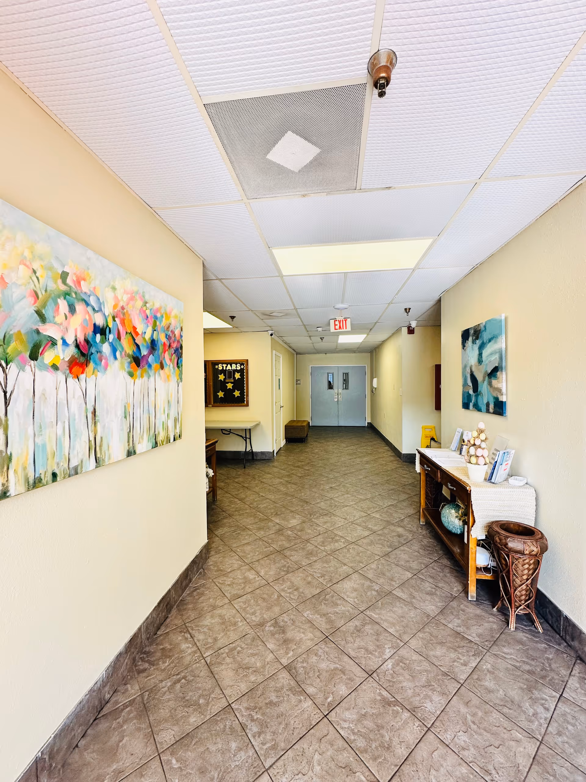 A clean, well-lit hallway in a senior living facility with tiled floors and beige walls. The hallway features colorful abstract paintings on both walls, a wooden console table with decorative items on the right, and a bulletin board labeled 'STARS' on the left. Double doors are visible at the end of the hallway with an exit sign above them.