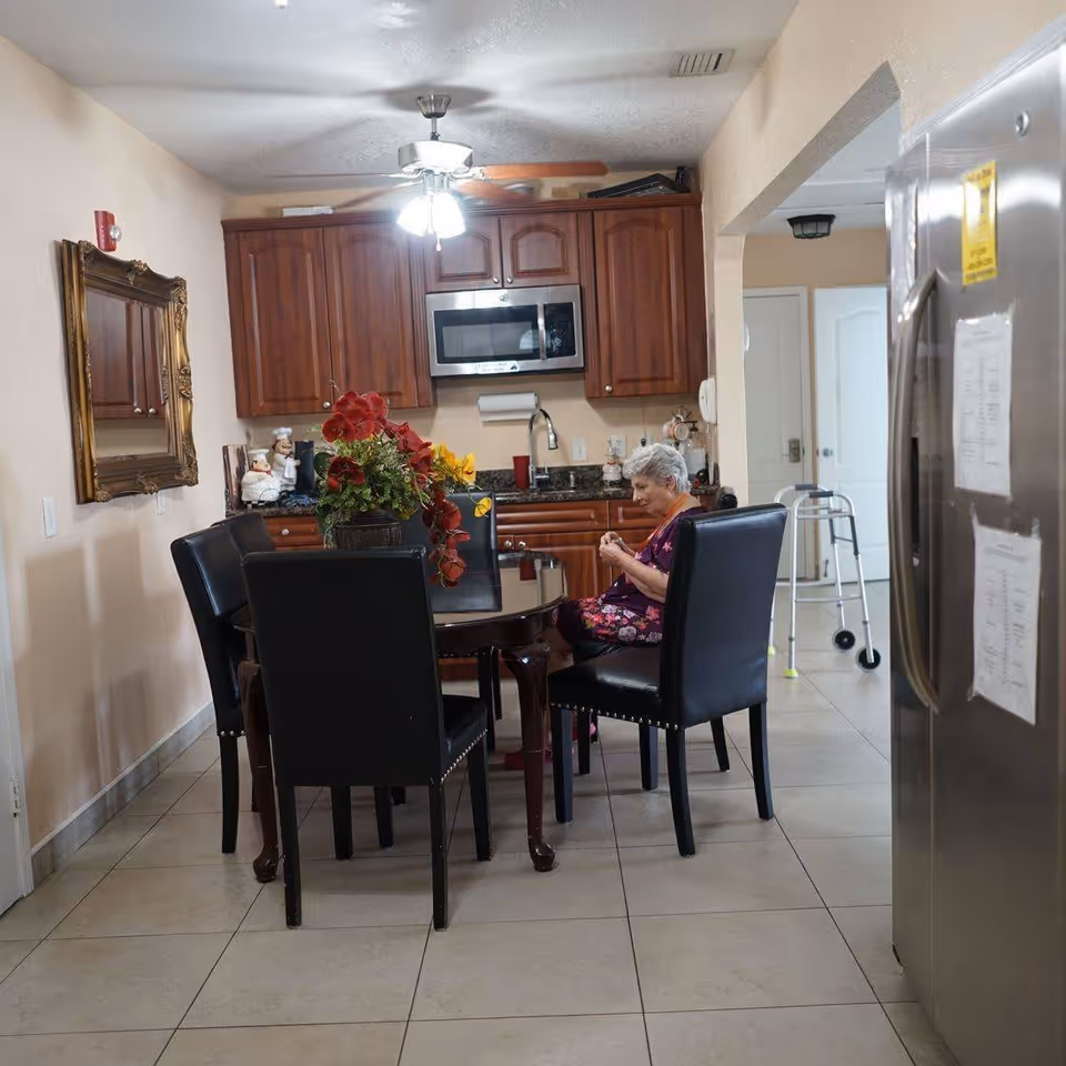 An elderly woman sitting at a round dining table in a kitchen area. The kitchen has wooden cabinets, a microwave, and a ceiling fan with lights. There is a large floral centerpiece on the table and black leather chairs around it. A walker is visible in the background near a doorway, and a stainless steel refrigerator is on the right side of the image.