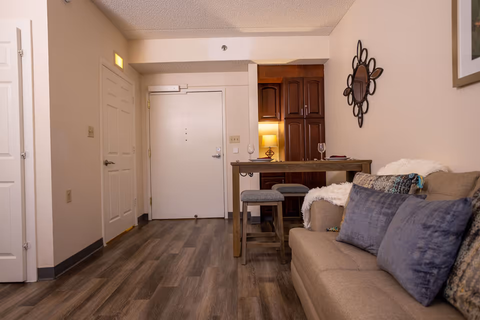 Interior view of a senior living facility apartment showing the entrance door, a small dining table with two stools, a beige sofa with multiple cushions, a decorative wall mirror, and wooden flooring.