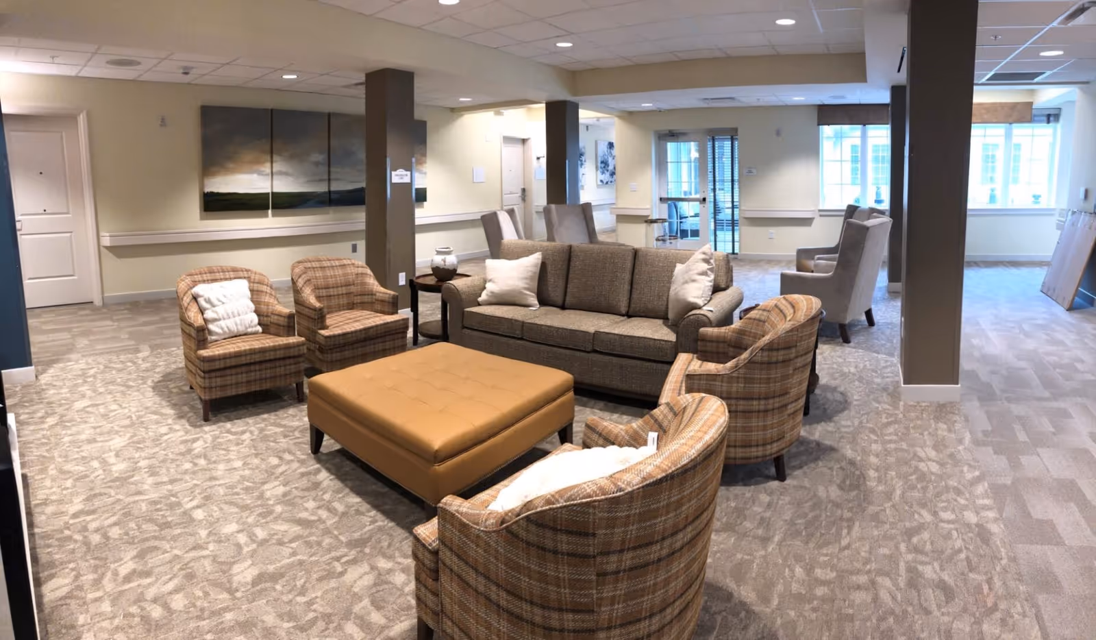 A spacious and well-lit living room area in a senior living facility with a beige sofa, four plaid armchairs, a large tan ottoman, and two high-back chairs near the windows. The room has carpeted floors, beige walls, and several columns. There is a triptych landscape painting on the wall and large windows letting in natural light.
