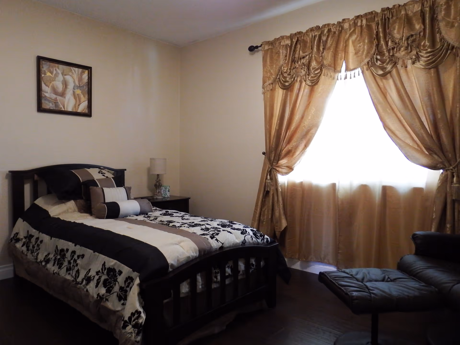 A neatly decorated bedroom with a bed, nightstand and lamp, gold curtains covering a bright window, and a leather lounge chair.