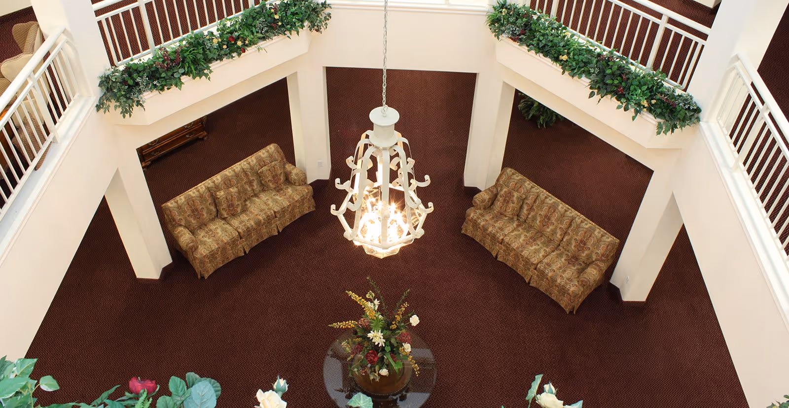View from above of a spacious indoor common area with two patterned sofas facing each other on a dark red carpet. A large white chandelier hangs in the center, and there is a round glass table with a floral arrangement below it. The area is surrounded by white railings with green plants along the edges.