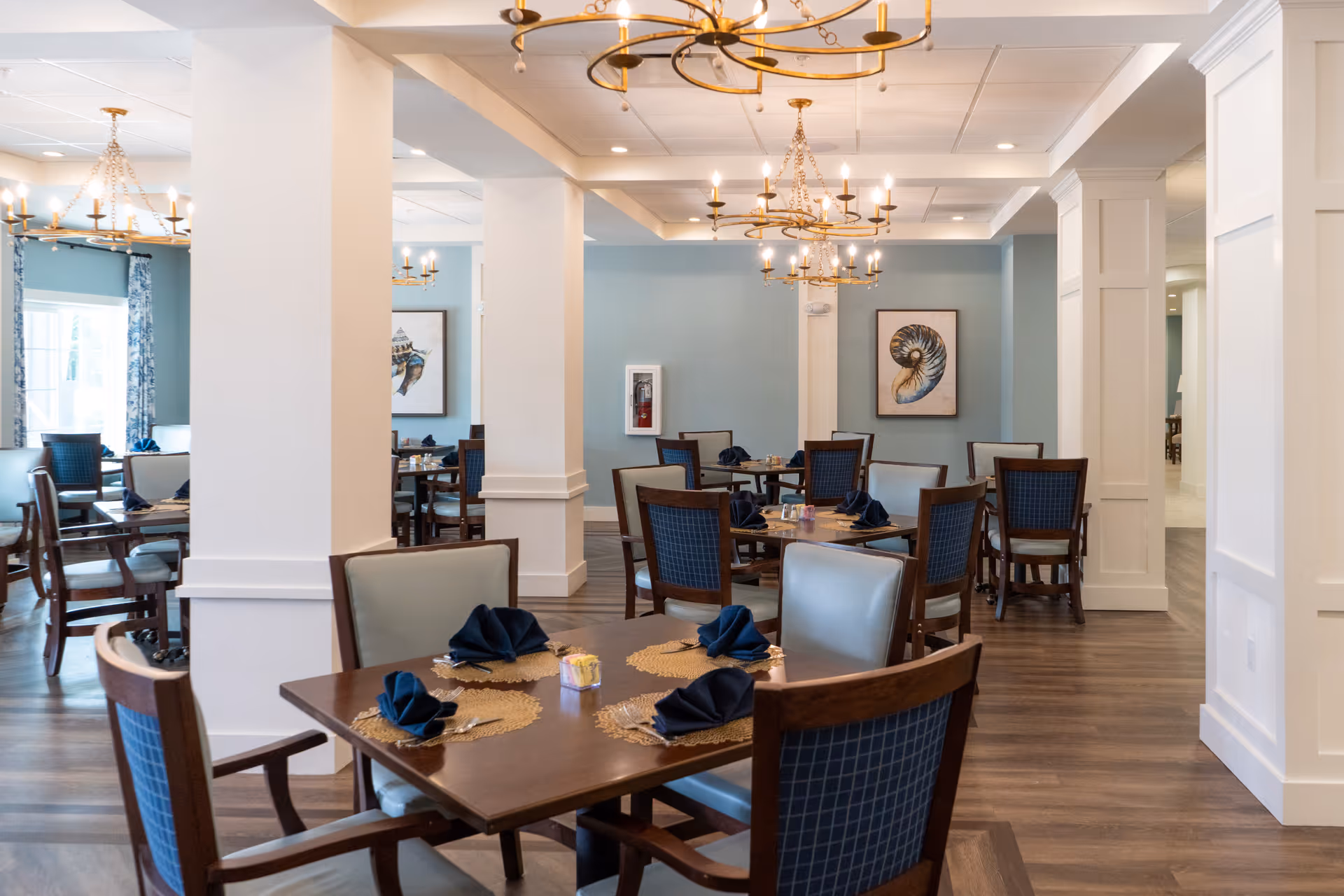 Spacious dining room with several set wooden tables and chairs, chandeliers, and soft blue walls.