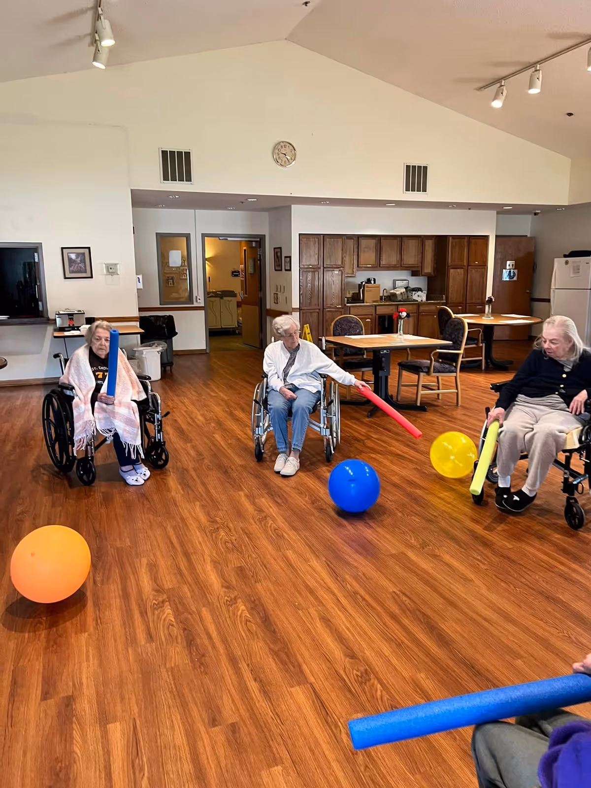 Three elderly women in wheelchairs participating in a group activity in a spacious room with wooden flooring. They are using colorful foam noodles to hit large balloons. The room has tables and chairs, a kitchenette area with wooden cabinets, and a clock on the wall.