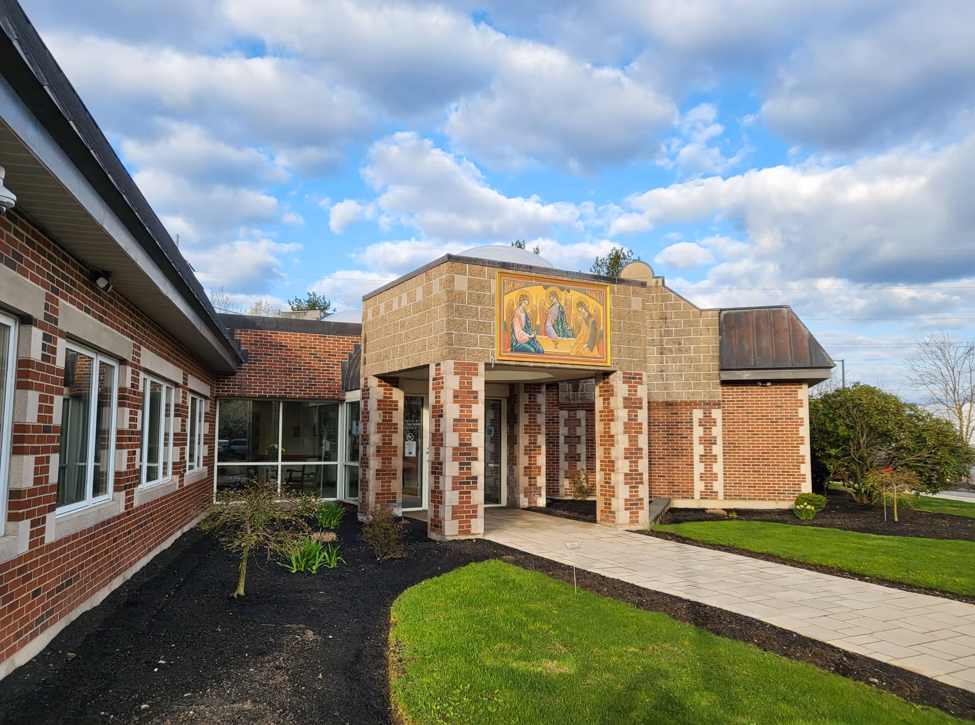 Exterior view of Holy Trinity Nursing and Rehabilitation Center showing a brick building with a covered entrance supported by brick columns. Above the entrance is a religious mural. The building is surrounded by green grass, small bushes, and a paved walkway under a partly cloudy sky.