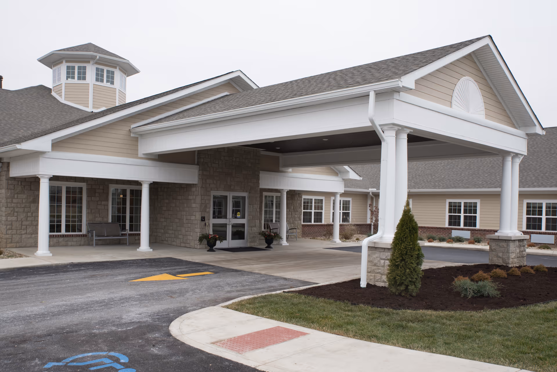 Front entrance of a senior living facility showing a covered porte-cochere with white columns, stone and siding facade, and a landscaped driveway.