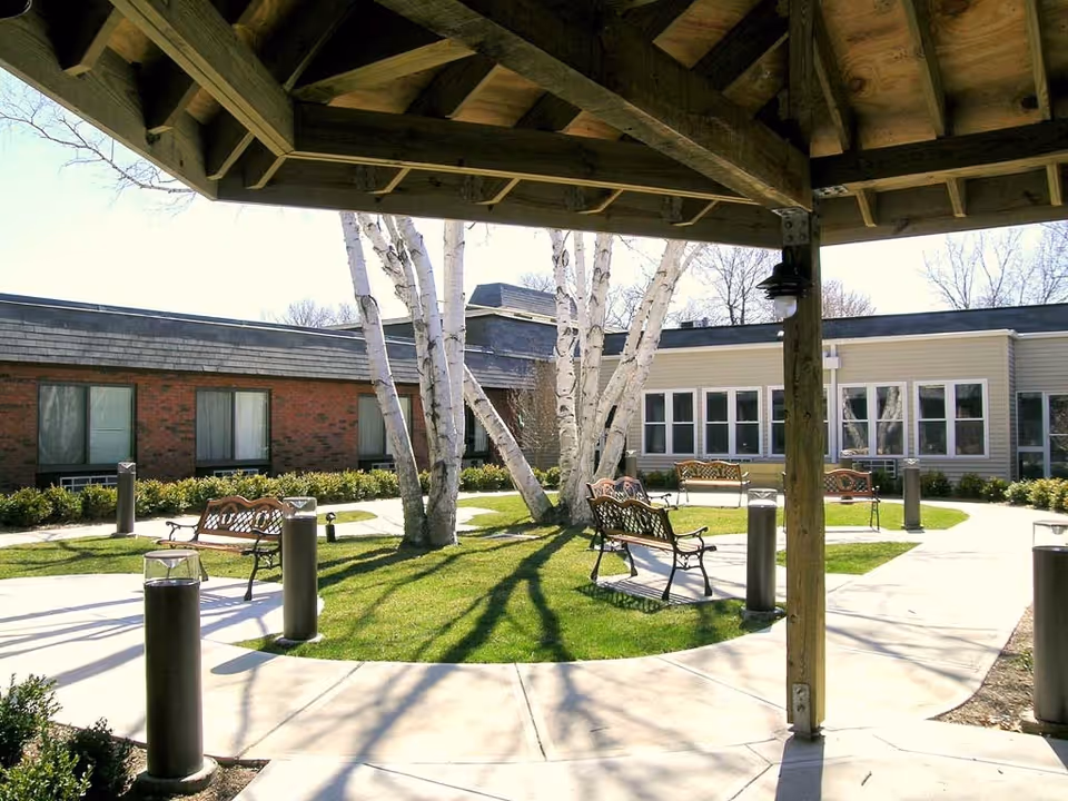 View from under a wooden gazebo of a sunny courtyard with benches, birch trees, and a surrounding single-story care building.