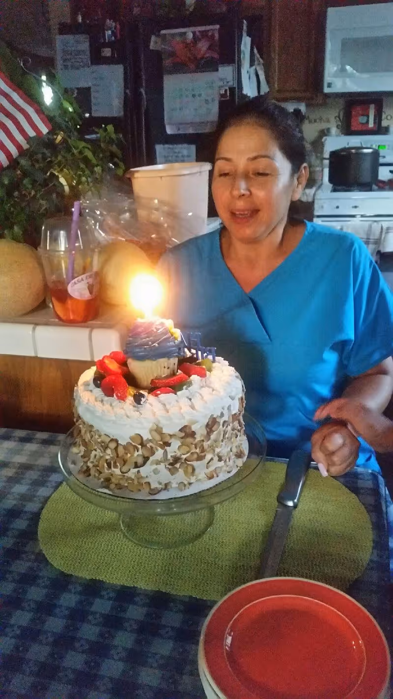 A woman in blue scrubs is sitting at a table with a birthday cake in front of her. The cake is decorated with whipped cream, nuts, strawberries, and a cupcake with a lit candle on top. The woman is holding someone's hand and appears to be making a wish or about to blow out the candle. The setting is a kitchen with various items on the counter and a stove in the background.