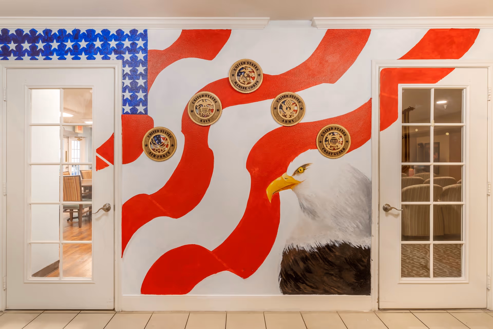 Interior wall mural featuring a large American flag with red and white stripes and a blue field with white stars. Five circular plaques representing the United States Air Force, Navy, Marine Corps, Army, and Coast Guard are mounted on the wall. A detailed painting of a bald eagle's head is on the lower right side of the mural. Two white doors with glass panes flank the mural, showing glimpses of rooms with chairs inside.