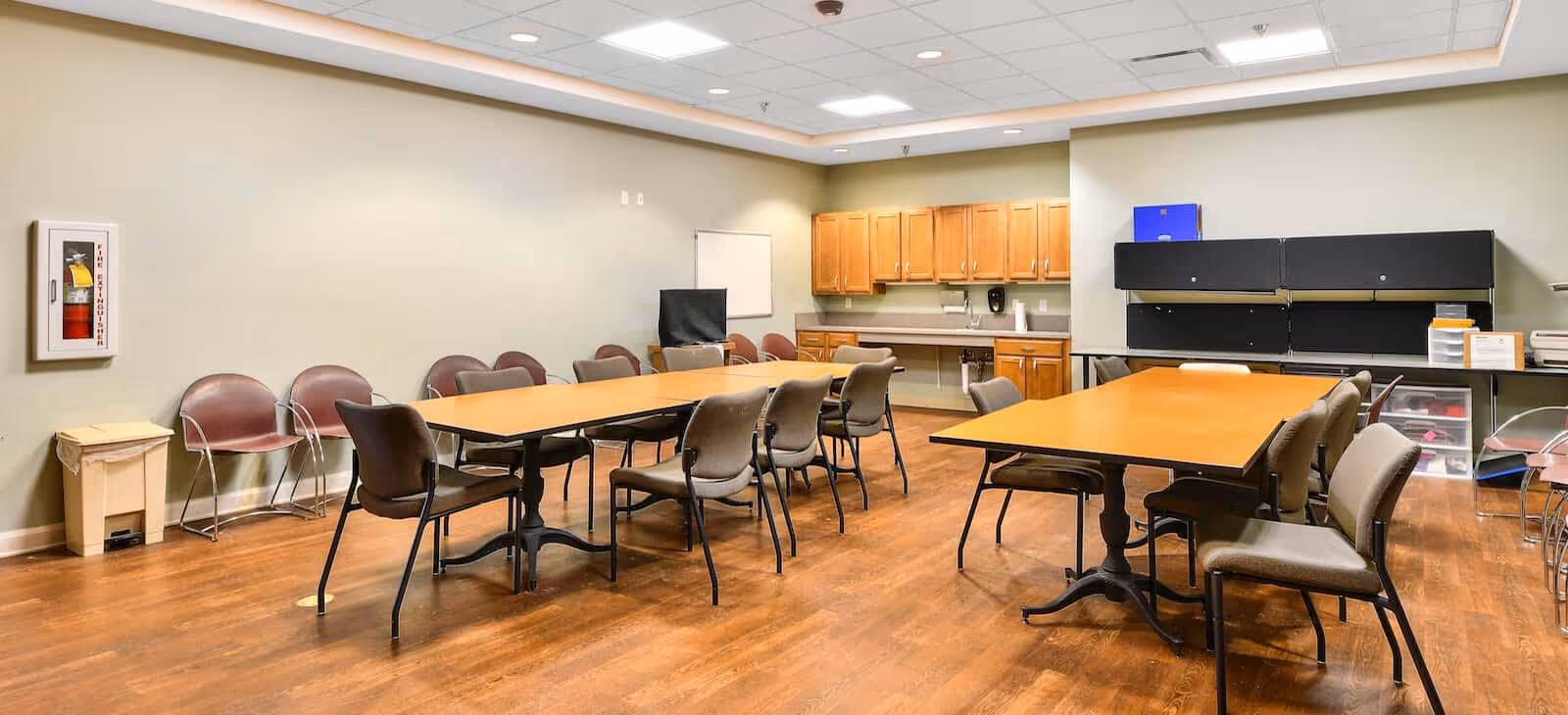 A spacious room with wooden flooring featuring two large rectangular tables surrounded by chairs. The walls are light-colored, and there are wooden cabinets and a countertop along the back wall. Additional chairs are lined up against the walls, and there is a fire extinguisher mounted on the left wall. The ceiling has recessed lighting.