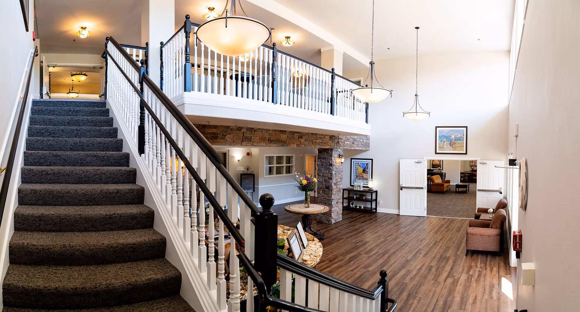 Interior view of a senior living facility showing a carpeted staircase with white and black railing leading to an upper floor. The area has wooden flooring, hanging pendant lights, a round table with a flower vase, framed artwork on the walls, and a seating area with chairs near double doors leading to another room.