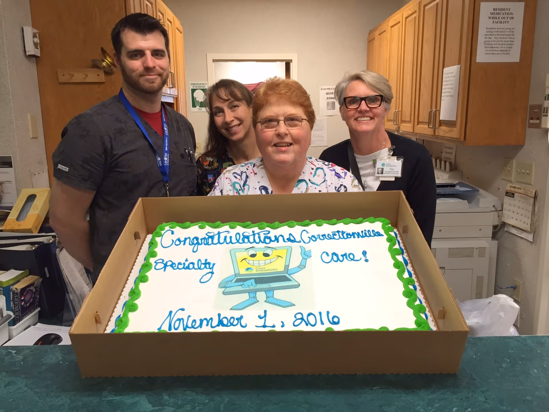 Four staff members standing behind a large congratulatory cake in a facility office area. The cake has green and blue icing and reads 'Congratulations Correctionville Specialty Care! November 1, 2016' with a cartoon laptop illustration. The background shows wooden cabinets, office equipment, and posted signs.