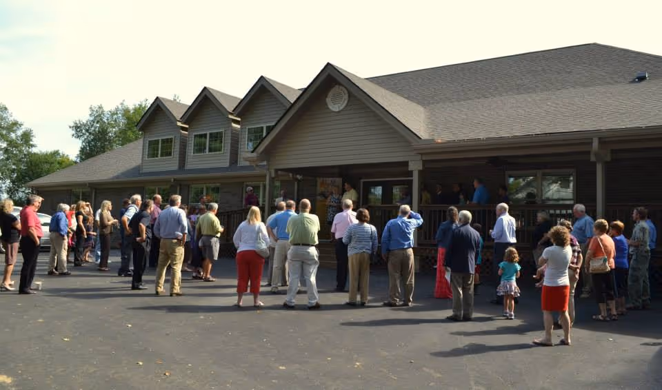 A group of people standing outside a large building with a porch, listening to a speaker. The building has multiple windows and a sloped roof, and the scene is set on a paved area with trees in the background.