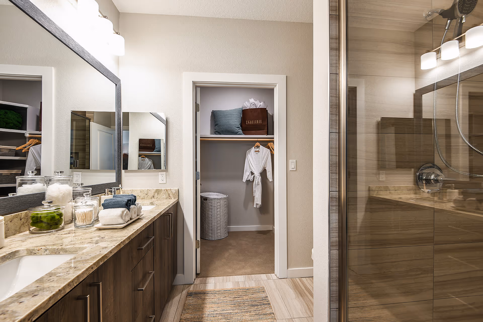 A modern bathroom with a double sink vanity featuring a granite countertop and wooden cabinets. On the countertop are glass jars with cotton balls, cotton swabs, and green decorative items, along with neatly folded white and blue towels. A large mirror with a dark frame is mounted above the sinks, and a light fixture with three lights is above the mirror. To the right is a glass-enclosed shower with beige tile walls. In the center background, there is an open doorway leading to a walk-in closet with a white robe hanging on a wooden hanger, a wicker laundry basket, a blue pillow, and a brown shopping bag on the shelf.
