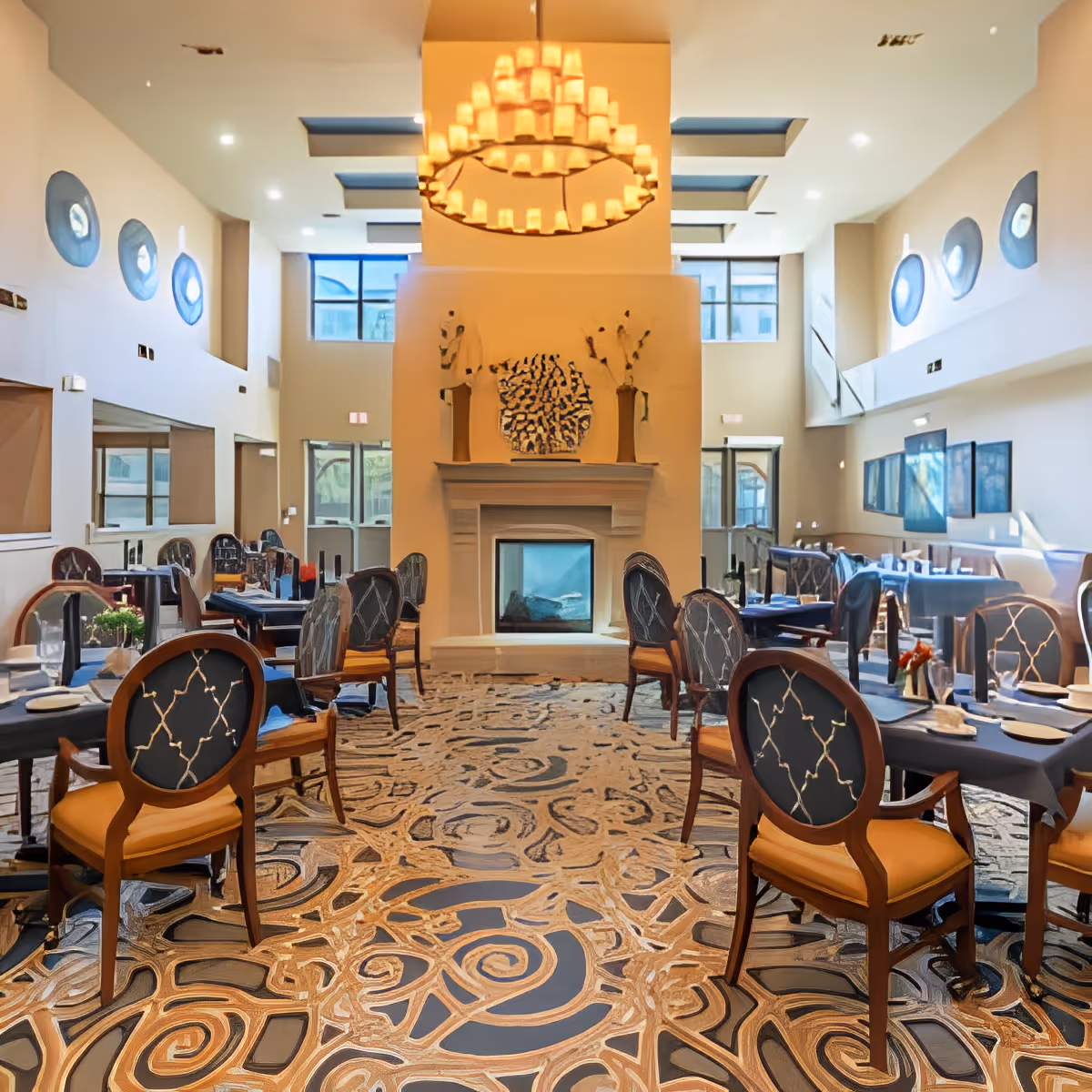 Double-height dining room with set tables and round-backed chairs, a central fireplace, and a large chandelier.