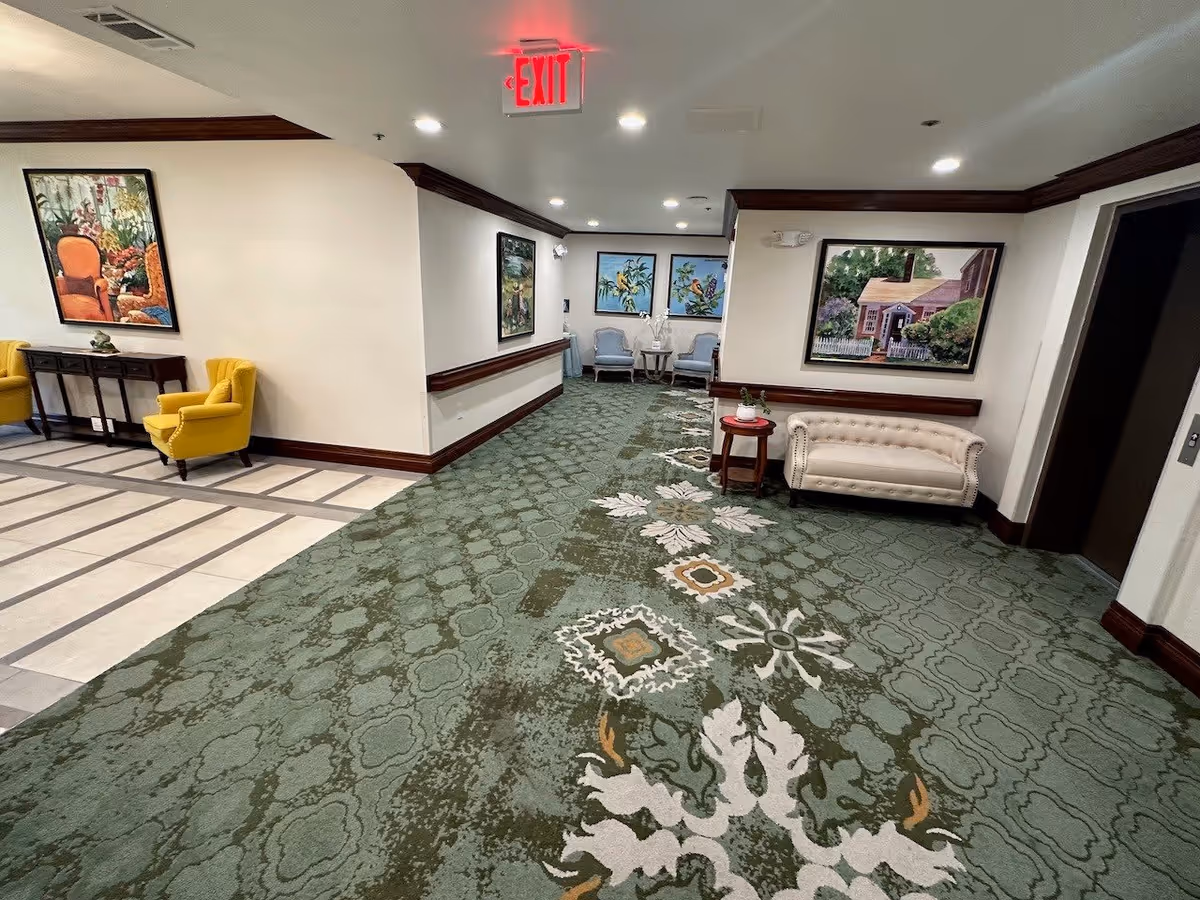 Interior hallway of a senior living facility with green patterned carpet and cream walls. The hallway features various seating areas including yellow armchairs, blue chairs, and a beige tufted sofa. Several framed paintings depicting nature and houses hang on the walls. An exit sign is visible on the ceiling.