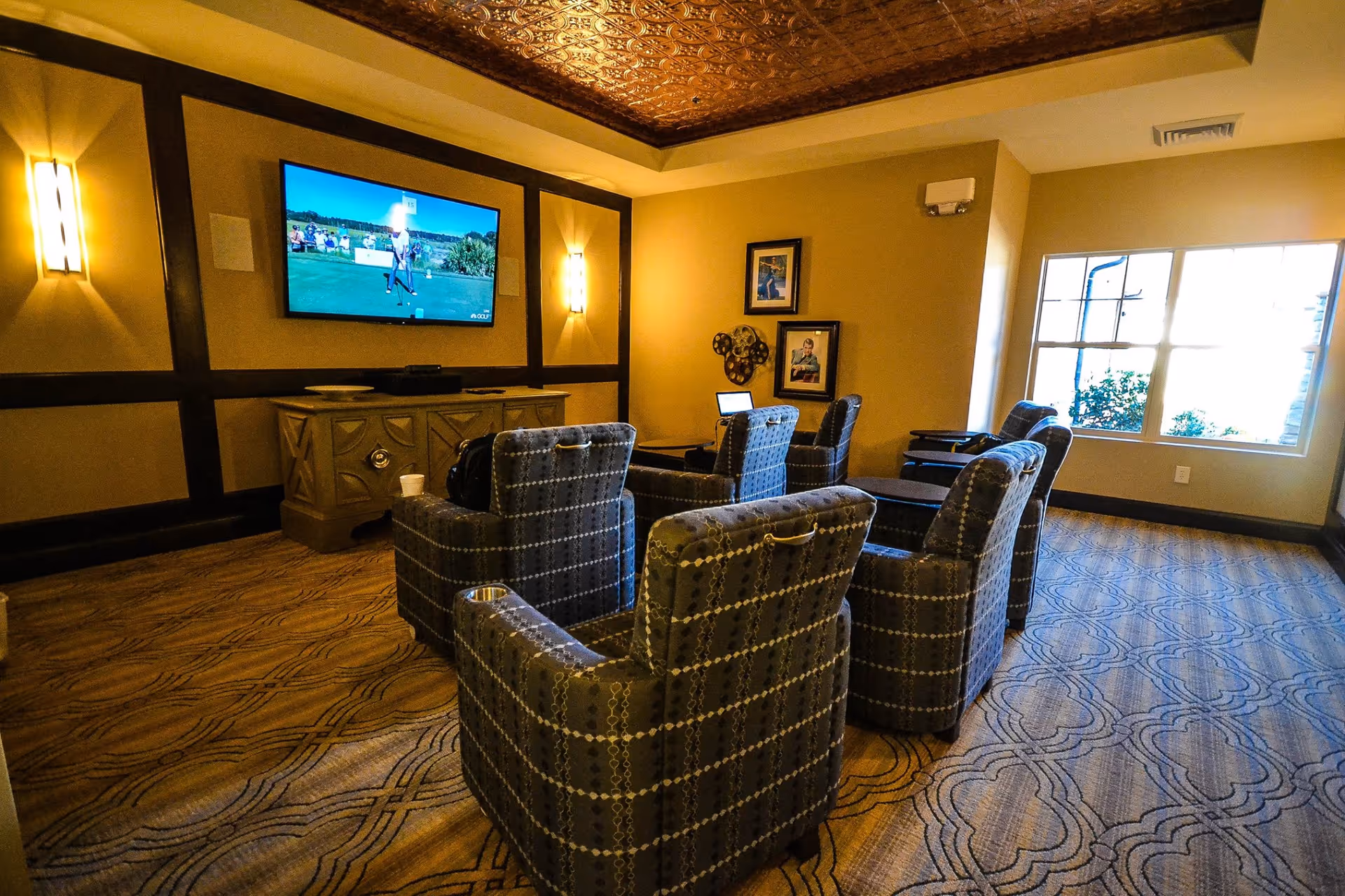A cozy media lounge with patterned armchairs arranged facing a wall-mounted TV and a window to the right.