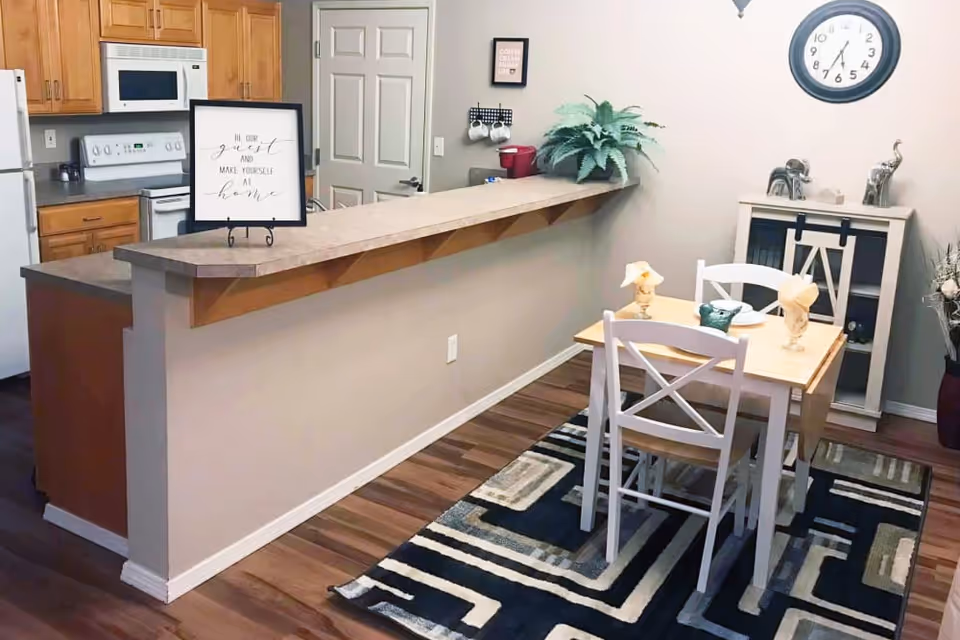 A cozy dining area and kitchen in a senior living facility. The dining area features a small wooden table with two white chairs, set with napkins and a small centerpiece on a patterned rug. The kitchen has wooden cabinets, a white refrigerator, stove, and microwave. A counter with a raised bar separates the kitchen from the dining area. Decorative items include a framed sign on the counter, a wall clock, a small cabinet with decorative elephants, and a potted plant on the counter.