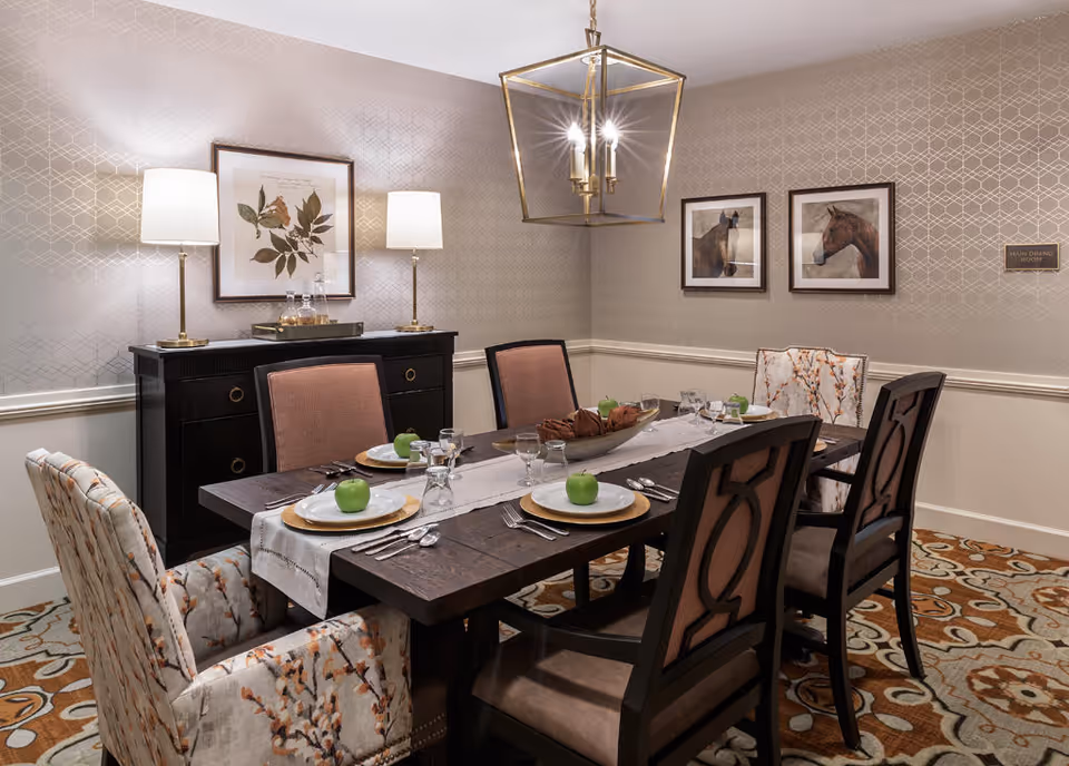 A well-decorated dining room with a dark wooden table set for six people. The table has white plates with green apples on each, silverware, glasses, and a centerpiece bowl with decorative items. The room features patterned wallpaper, two framed pictures of horses on one wall, a framed botanical print on another wall, two table lamps on a black sideboard, and a geometric chandelier hanging above the table. The carpet has an intricate floral pattern in shades of brown, beige, and blue.