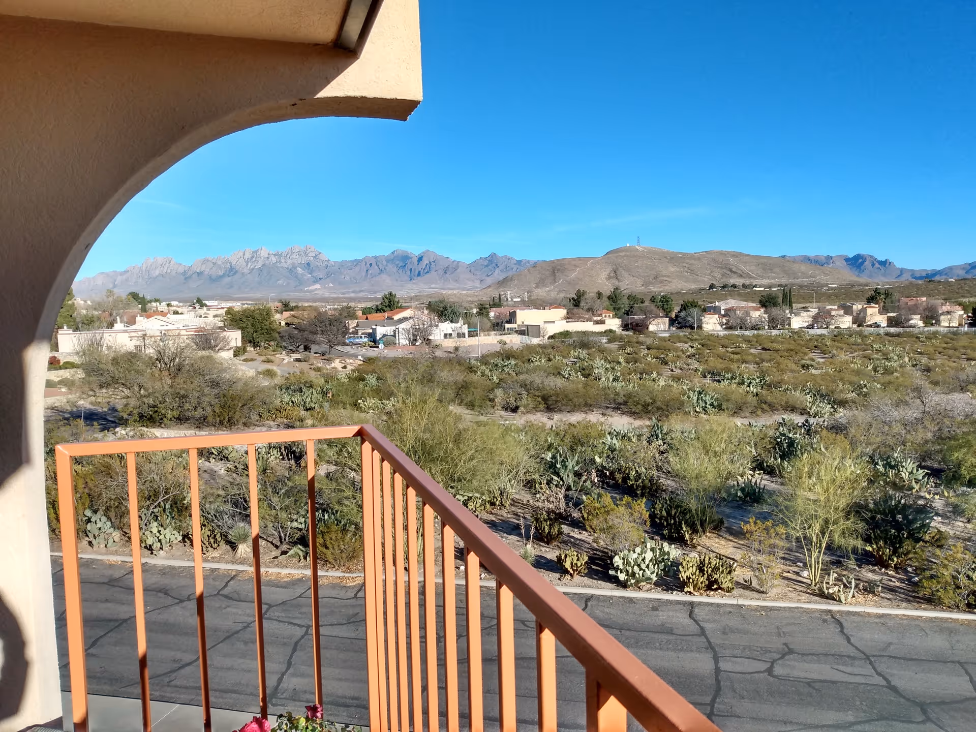 View from a balcony with an orange railing and archway overlooking desert scrub, nearby houses and distant mountains under a clear blue sky.