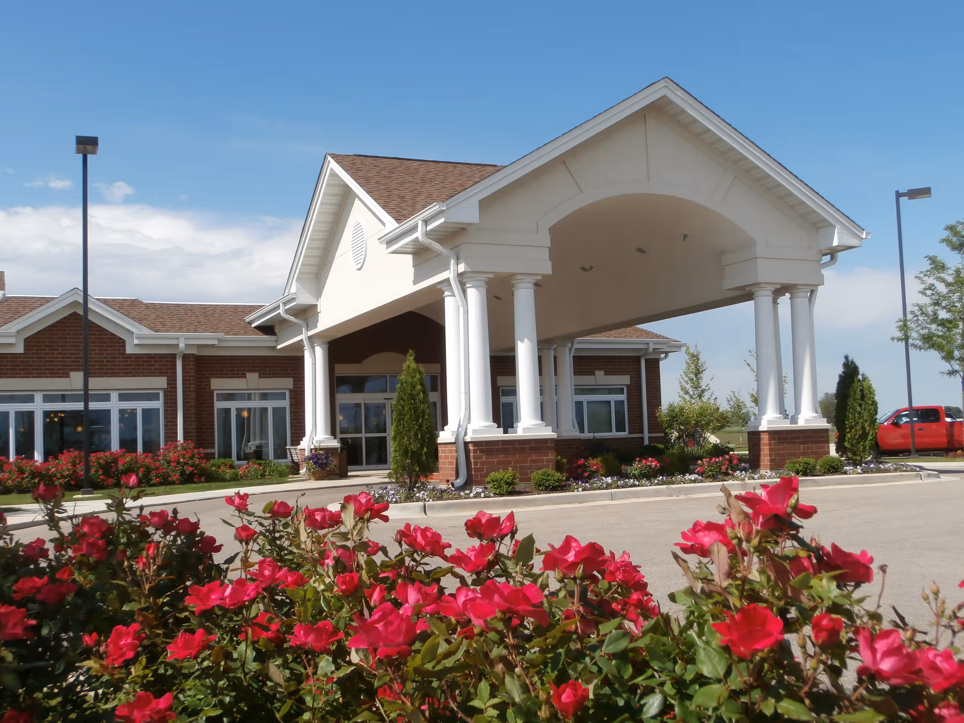 Entrance of a senior living facility with a large covered driveway supported by white columns. The building has a brick exterior with large windows. In the foreground, there are vibrant red flowers and green shrubs. A red truck is parked to the right side of the building under a clear blue sky.
