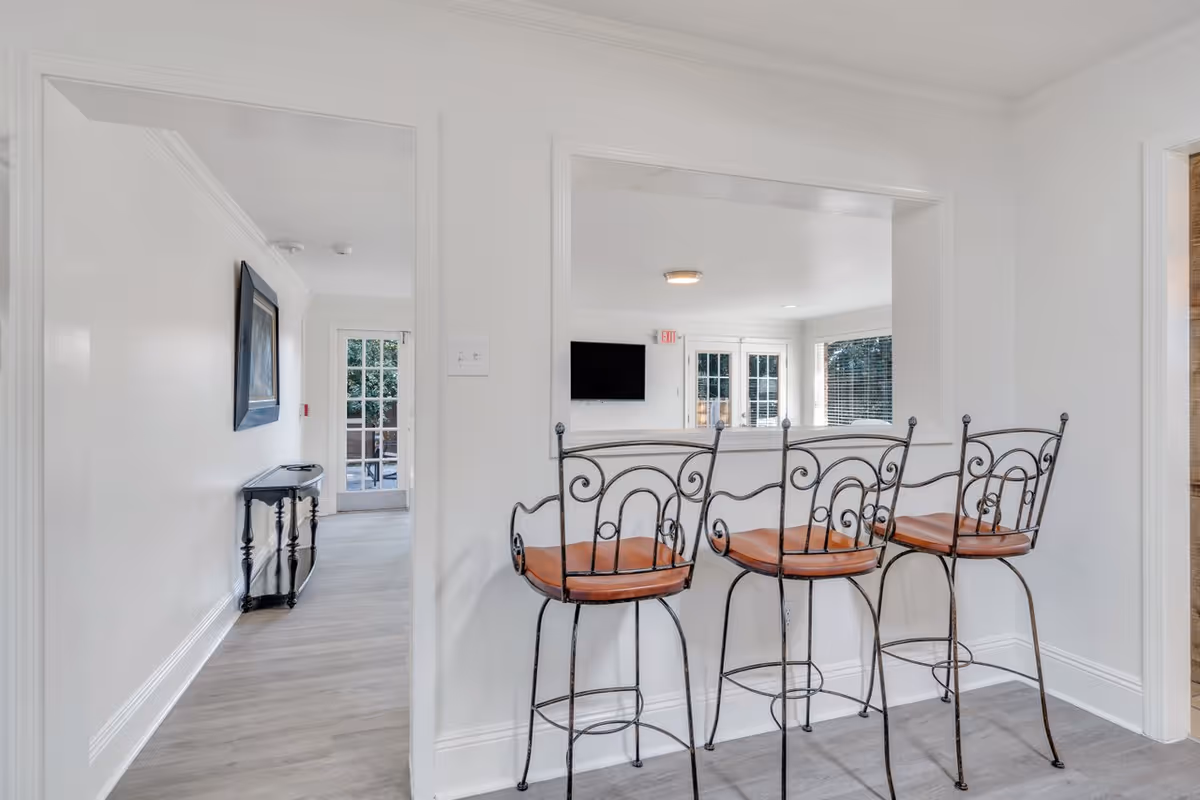 Interior view of a senior living facility showing a counter with three wrought iron bar stools with brown seats. The counter opens to a bright room with large windows and a wall-mounted TV. To the left, there is a hallway with a small black console table and a framed picture on the wall. The walls are white and the floor is light-colored wood.