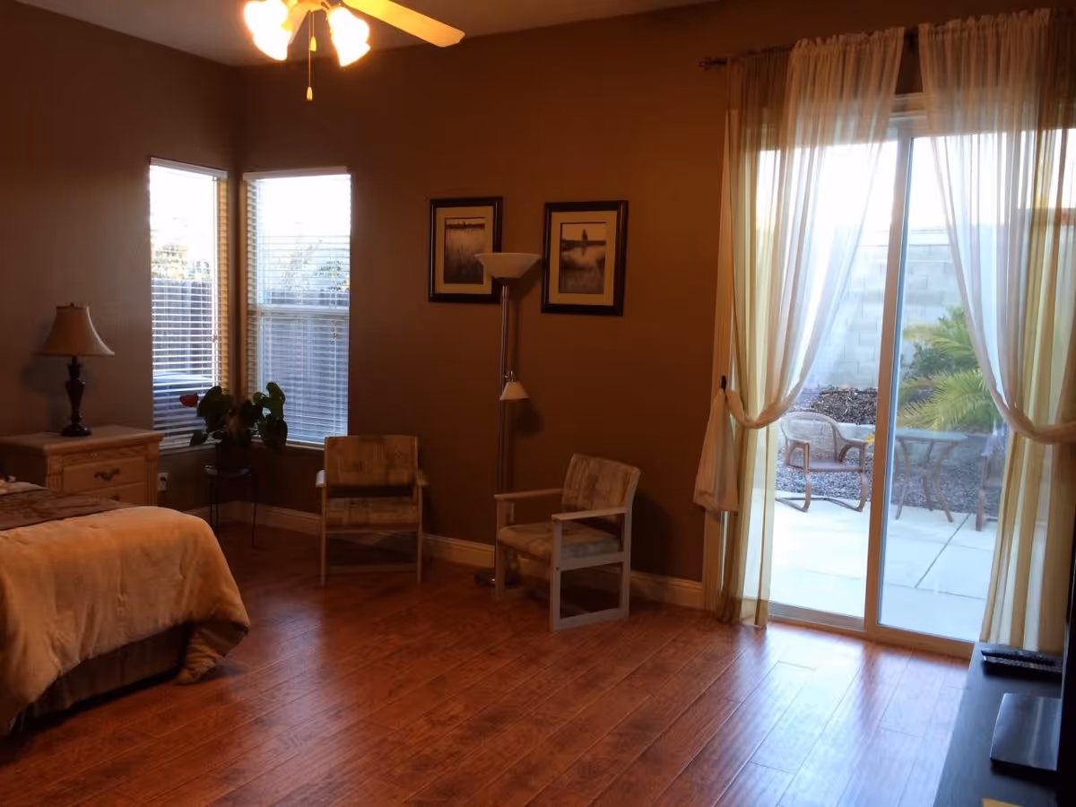 A cozy bedroom with wooden flooring, a bed with beige bedding, a nightstand with a lamp, two armchairs, a floor lamp, two framed pictures on the wall, and large windows with blinds. There is a sliding glass door with sheer curtains leading to an outdoor patio area with chairs and a table.