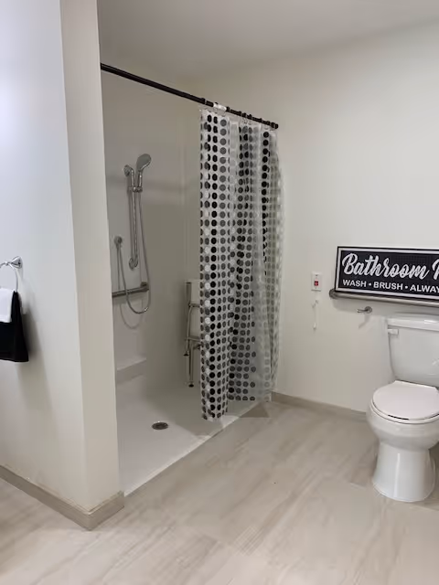 A clean, modern bathroom featuring a walk-in shower with a black and white polka dot shower curtain, a white toilet, and a wall sign that reads 'Bathroom Rules: Wash, Brush, Always Smile'. The floor is light-colored tile and the walls are painted white.