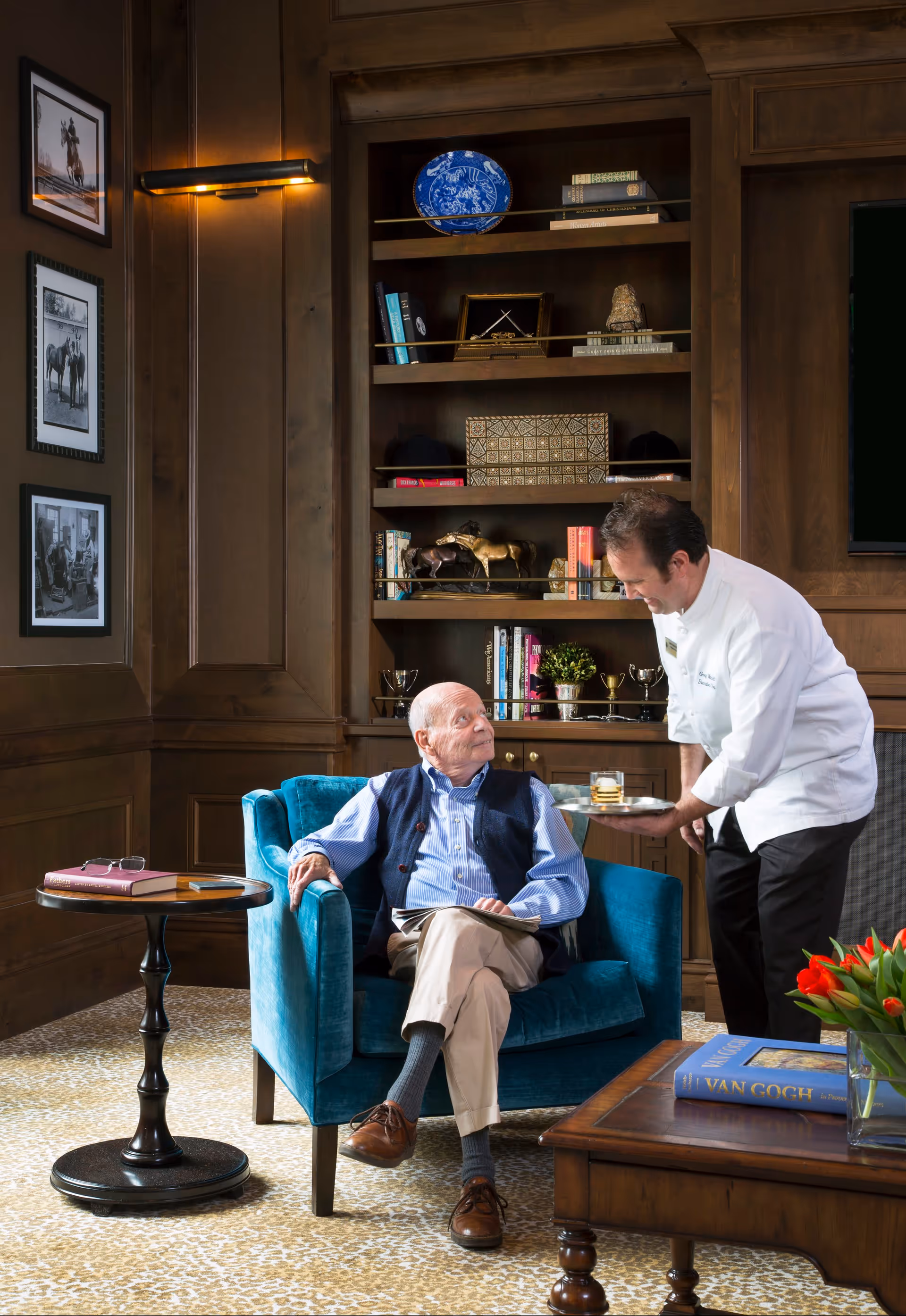 An elderly man sitting in a blue armchair in a wood-paneled room with built-in bookshelves. A staff member in a white uniform is serving him a drink on a tray. The room has framed black and white photos on the wall, a small round side table with a book and glasses, and a coffee table with a book titled 'Van Gogh' and a vase of red tulips.