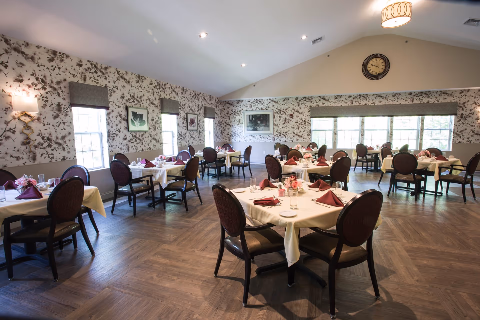A spacious dining room with multiple round tables covered with white tablecloths and set with burgundy napkins, glasses, and small floral centerpieces. The room has large windows with gray valances, floral wallpaper, wooden flooring, and a clock on the far wall under a vaulted ceiling with recessed lighting.