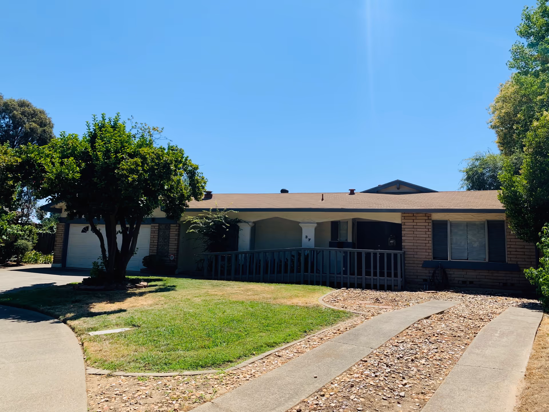 Single-story residential building with a sloped roof, a garage on the left, a front porch with a ramp, and a lawn with trees and shrubs under a clear blue sky.
