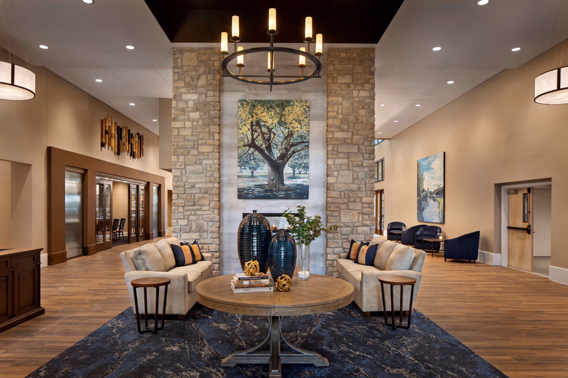A spacious and elegant living room area in Stevenson Oaks featuring two beige sofas with decorative pillows facing each other across a round wooden table adorned with vases, books, and decorative objects. The room has a stone pillar with a large painting of a tree, wooden flooring, modern lighting fixtures, and additional seating with blue chairs along the walls.
