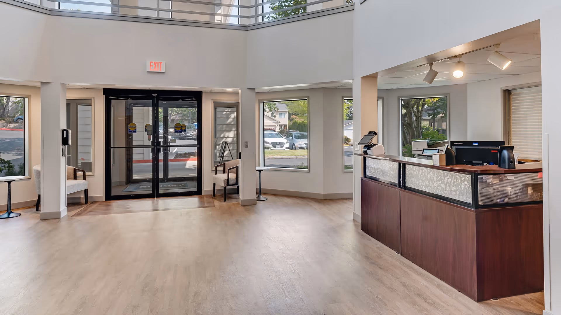 Bright and spacious lobby area with large windows, a double glass entrance door, seating areas with chairs and small tables, and a reception desk with computer equipment under a ceiling with recessed lighting.