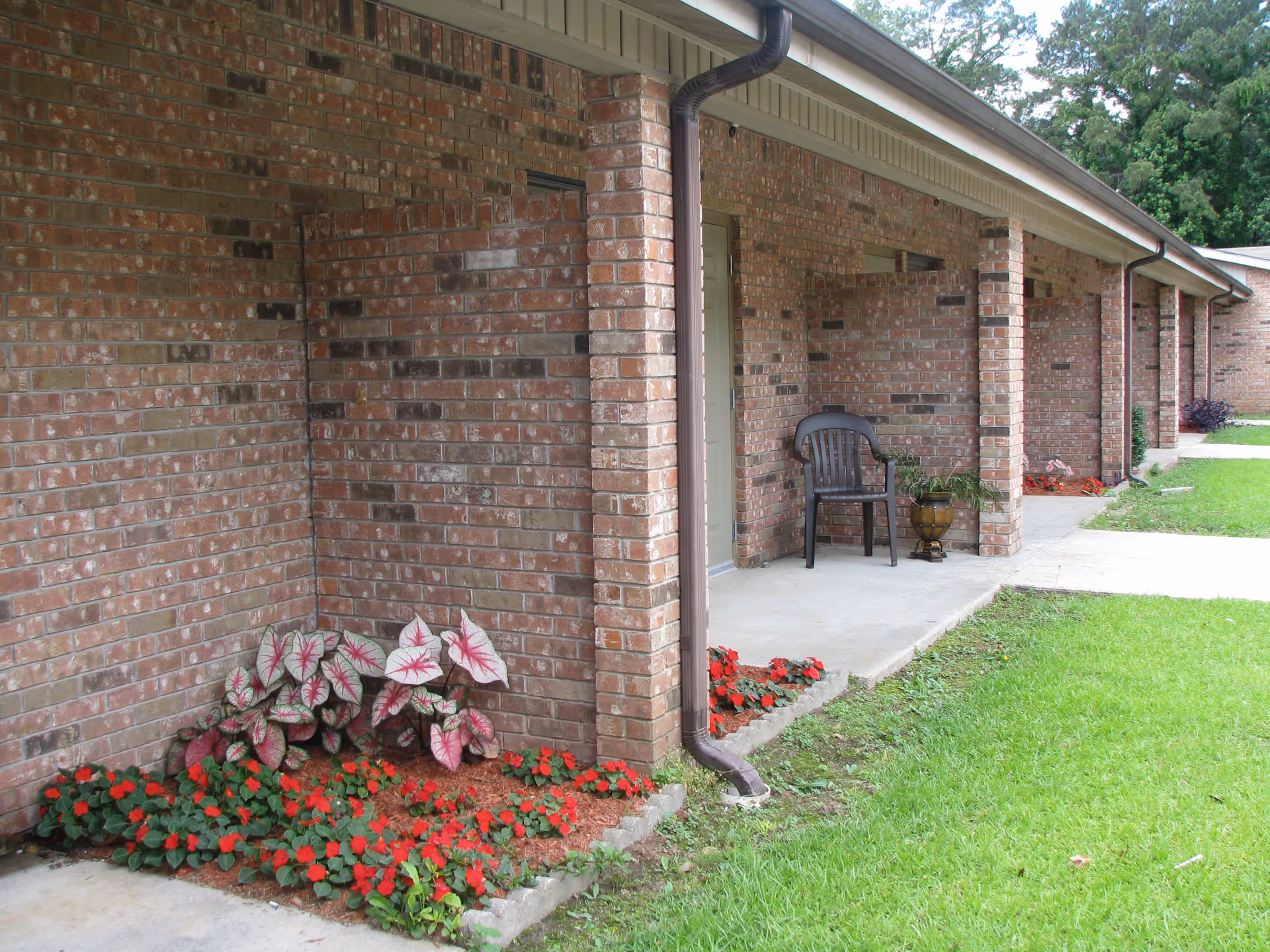 Exterior view of a brick building with a covered walkway. There are flower beds with red and green plants along the building, a black plastic chair, and a potted plant near the entrance. The area is surrounded by green grass and trees in the background.