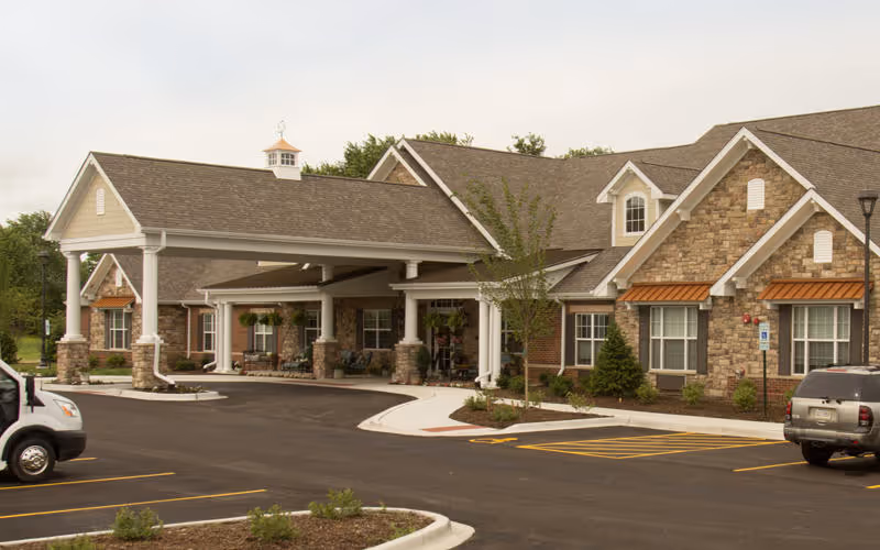 Exterior view of a senior living facility building with stone and brick facade, a covered entrance supported by white columns, and a parking lot with vehicles in front.