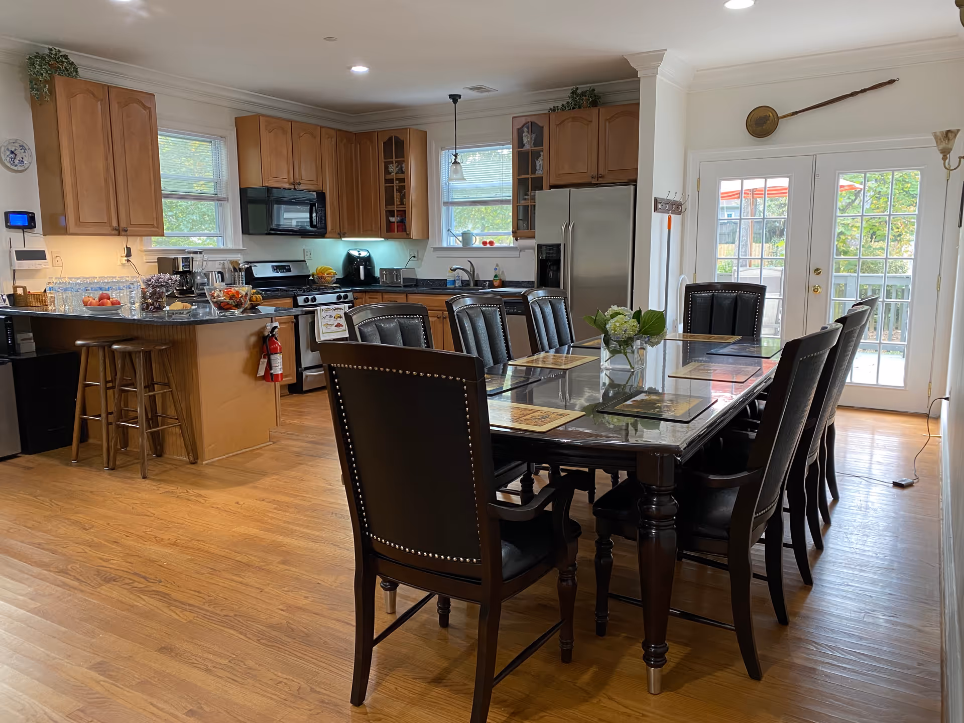 A spacious kitchen and dining area with wooden floors and cabinetry. The kitchen features stainless steel appliances including a refrigerator and stove, a microwave, and a countertop with stools. The dining area has a large rectangular table with eight black leather chairs, placemats, and a small flower arrangement in the center. French doors lead to an outdoor space with visible greenery and red umbrellas.