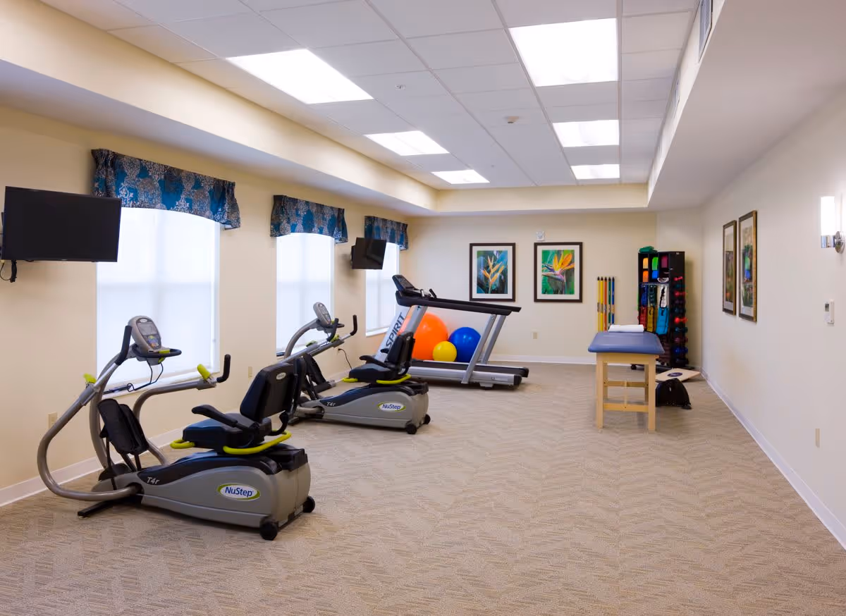 A well-lit exercise room with two NuStep recumbent cross trainers, a treadmill, colorful exercise balls, a massage or therapy table, and a rack with various exercise equipment. The room has beige walls, carpeted floor, three windows with blue patterned valances, and framed artwork on the walls.