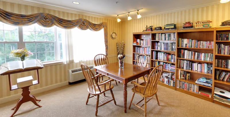 A cozy room with a wooden table and four wooden chairs arranged around it. There is a vase with decorative sticks on the table. The room has beige striped wallpaper and carpeted floor. A large window with patterned valance curtains lets in natural light. A wooden bookshelf filled with books and model trains on top lines one wall. A wooden stand holds an open book and a small flower arrangement near the window.