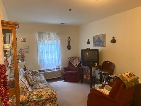 A cozy living room with floral patterned sofa, two red armchairs, a small TV on a stand, a window with sheer curtains, and various wall decorations including a clock and framed pictures.