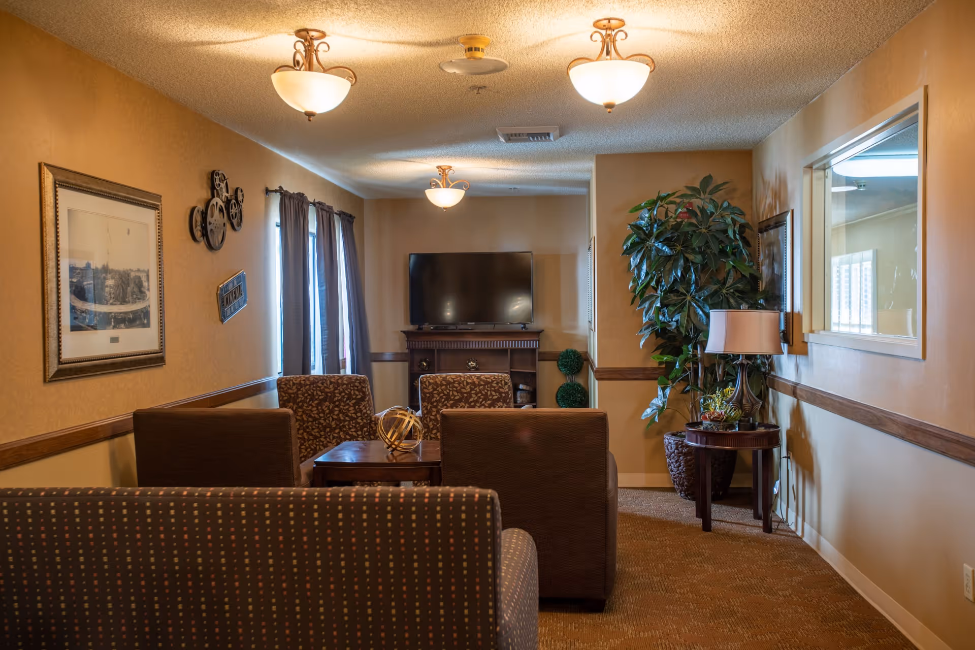 A cozy living room area in a senior living facility with patterned sofas and armchairs arranged around a wooden coffee table. The room features warm beige walls, three ceiling light fixtures, a flat-screen TV on a wooden stand, a large potted plant, a side table with a lamp, and framed artwork on the walls. There are windows with dark curtains letting in natural light.