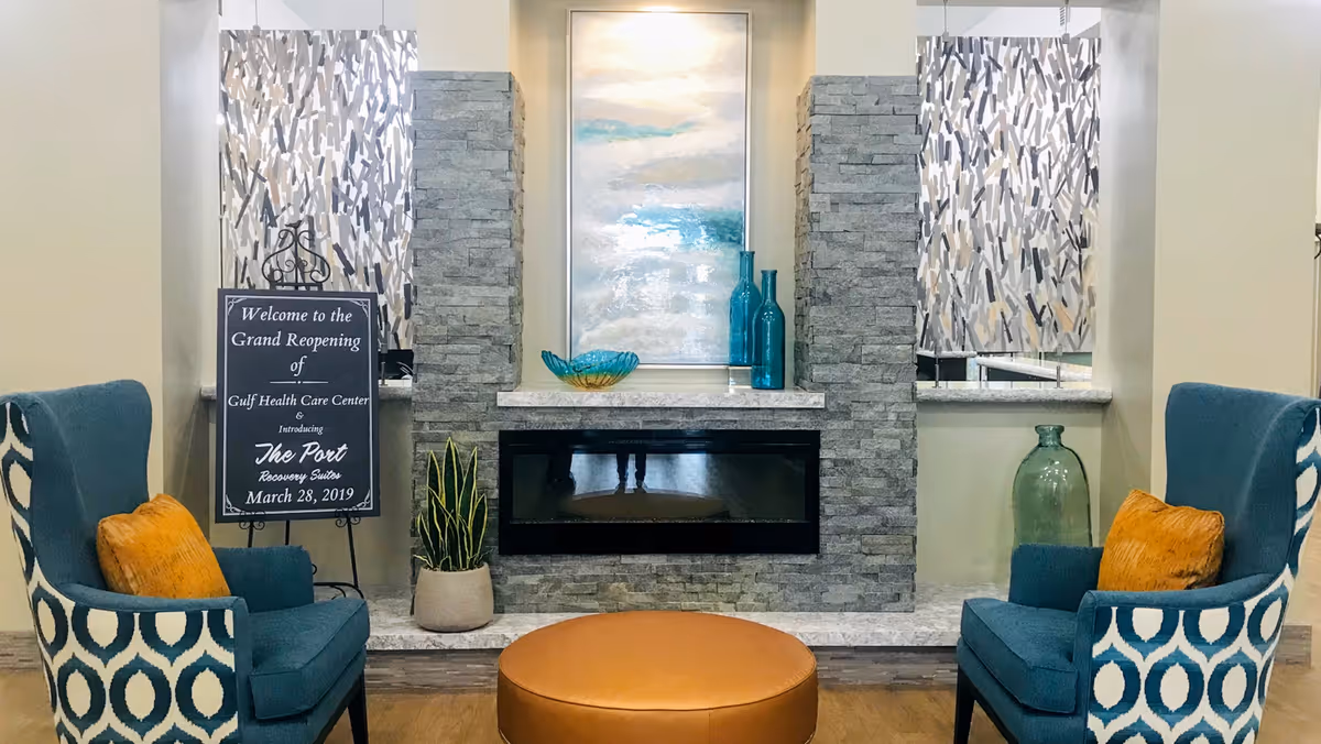 A cozy seating area with two blue patterned armchairs with orange cushions facing a modern stone fireplace. Above the fireplace is a large abstract painting with blue and white tones. On the mantel are decorative glass bottles and a bowl. A round brown ottoman is centered in front of the fireplace. To the left, a sign on an easel announces the grand reopening of Gulf Health Care Center and the introduction of The Post Recovery Suites on March 28, 2019. The room has light-colored walls and wood flooring.