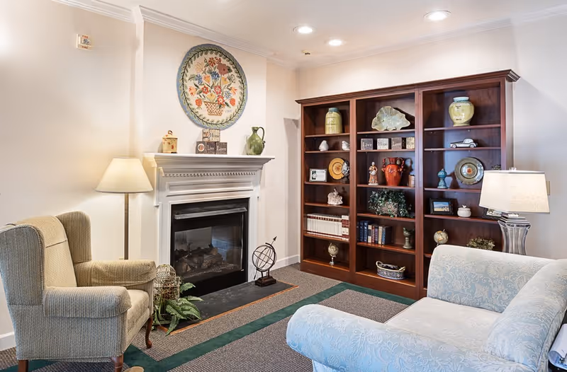 A cozy living room area featuring a beige armchair and a light blue patterned sofa. There is a white fireplace with decorative items on the mantel, including a green pitcher and a floral wall hanging above it. To the right, a large wooden bookshelf holds various decorative objects, books, and vases. Two lamps provide warm lighting in the room.